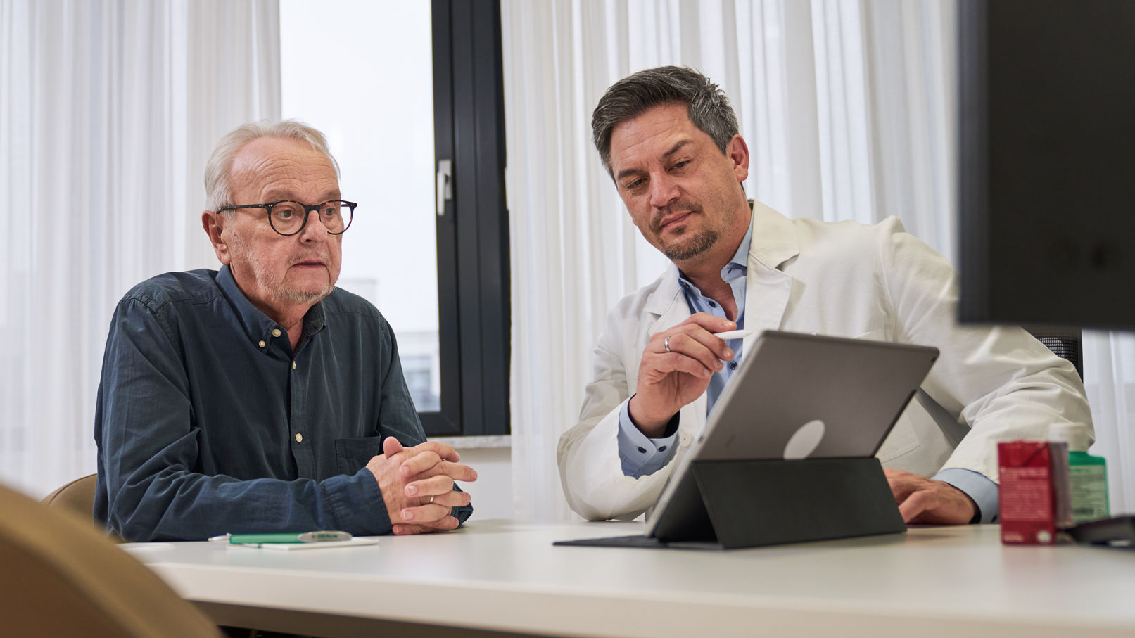 oncologist-consulting-cancer-patient-and-giving-advice-via-ipad-at-the-table-in-the-consultancy-room-about-adjuvant-nutritional-therapy-and-remune