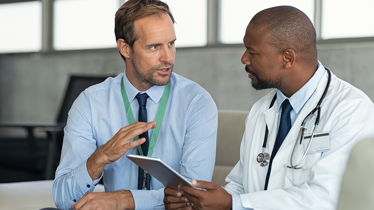 Représentant pharmaceutique discutant avec un médecin assis sur un divan. Praticien africain mature discutant des résultats de l’analyse avec un spécialiste tout en consultant le diagnostic sur une tablette numérique. Médecin en conversation avec un conseiller médical dans une chambre d’hôpital discutant du cas d’un patient.